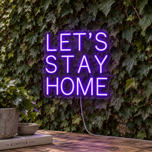 Let's Stay Home LED Neon Sign in purple, featuring three lines of block lettering against a foliage wall.