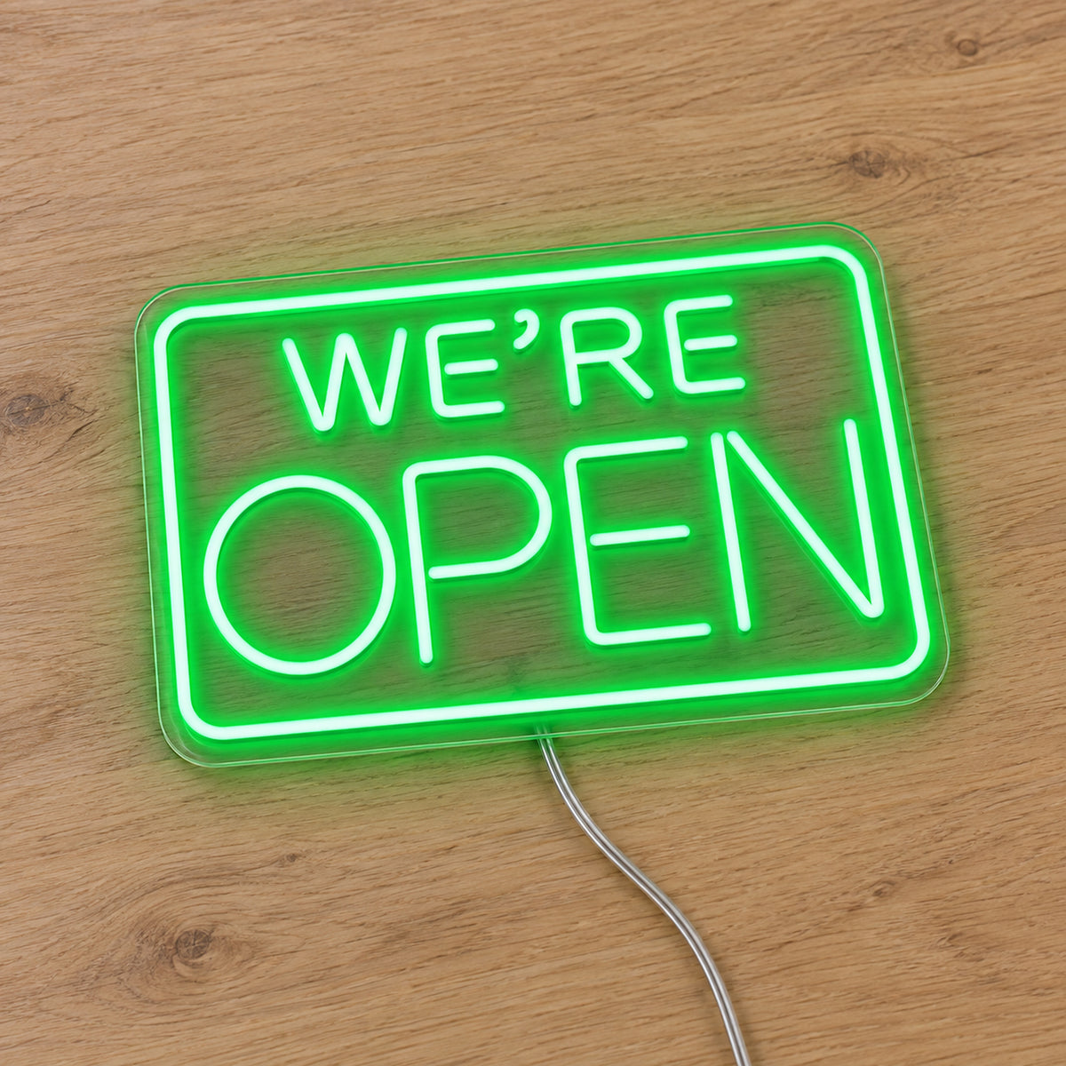 We Are Open LED Neon Sign in green, featuring We're Open lettering inside a rectangular border on a wooden surface.