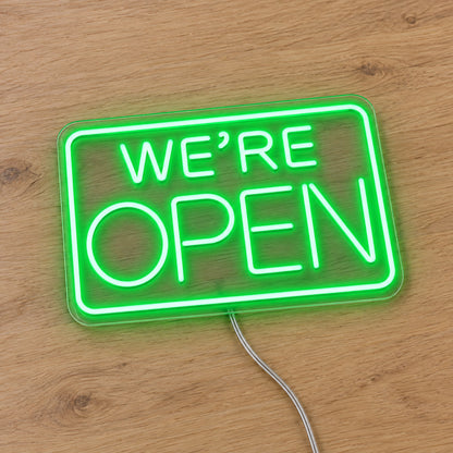 We Are Open LED Neon Sign in green, featuring We're Open lettering inside a rectangular border on a wooden surface.