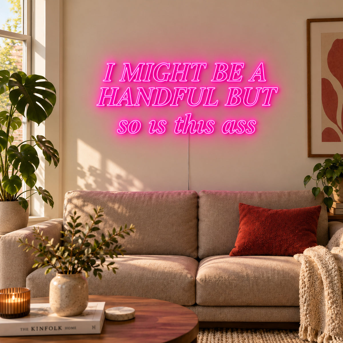 I Might Be A Handful But So Is This Ass Neon Sign glowing pink in compact lettering above a cream sofa.