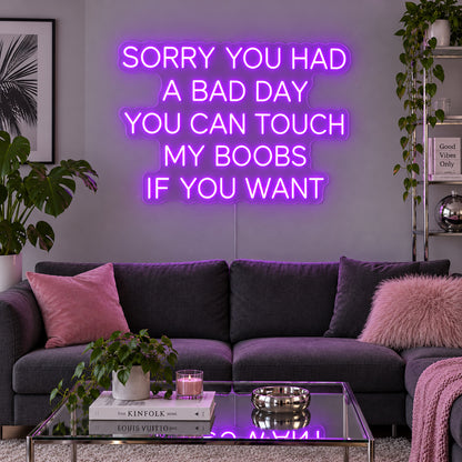 Sorry You Had A Bad Day You Can Touch My Boobs If You Want Neon Sign with purple lighting, displayed above a grey sofa with pink cushions.