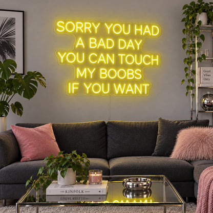 Sorry You Had A Bad Day You Can Touch My Boobs If You Want Neon Sign with yellow lighting, displayed above a grey sofa with pink cushions.