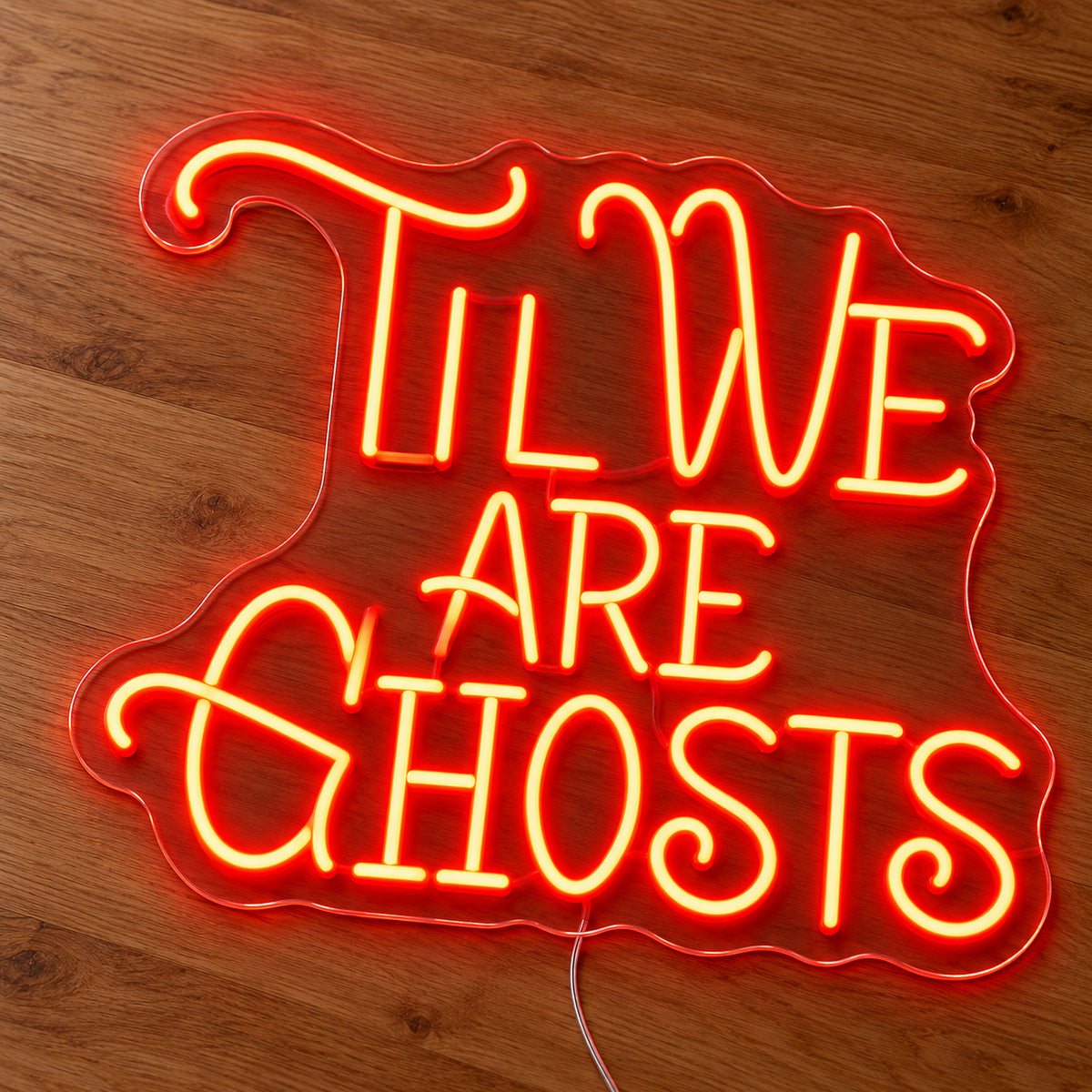 Til We Are Ghosts Ornate Neon Sign featuring Til We Are Ghosts wording in decorative orange lettering with curved flourishes, illuminated on clear acrylic lying on a natural oak wooden surface.